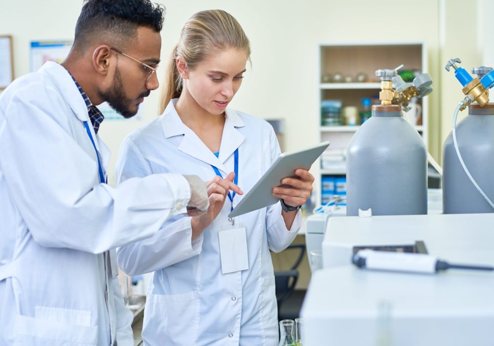 Serious thoughtful medical colleagues using digital tablet while viewing report together and discussing results standing by oxygen cylinders in laboratory
